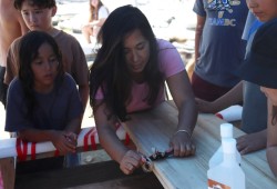 Nuu-chah-nulth youth gather around Lacy Kaheaku, learning how to carve a traditional wooden surf board and of the Indigenous roots sport.